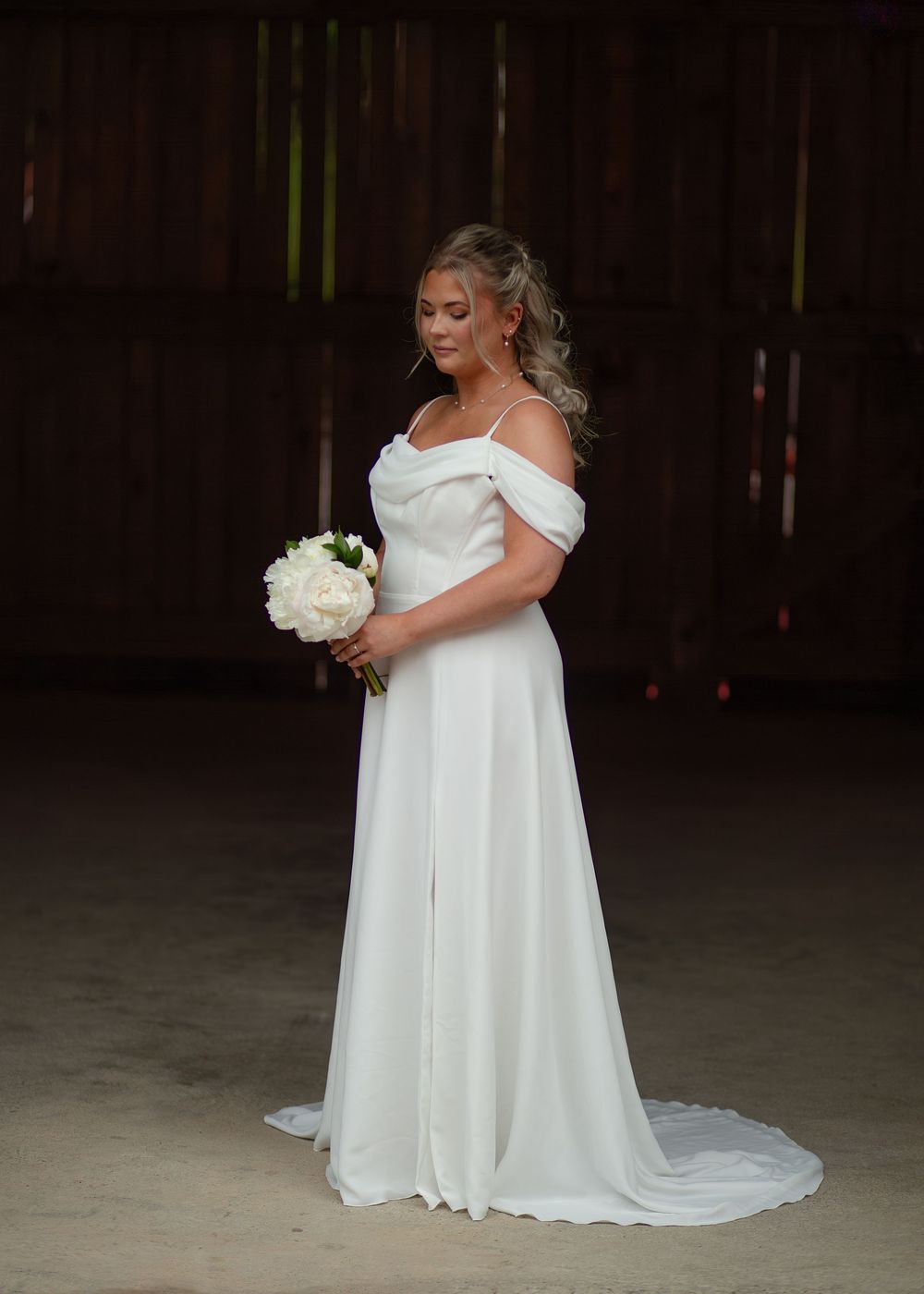 Bride in elegant off-shoulder white wedding gown holding white peony bouquet in rustic barn setting.