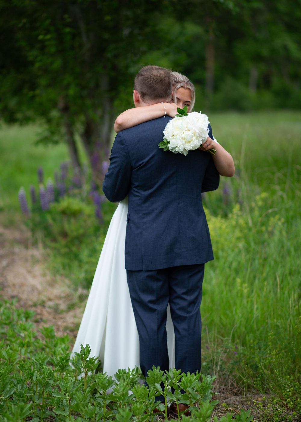 Bride and groom embracing in a lush green outdoor setting, bride holding white bouquet.