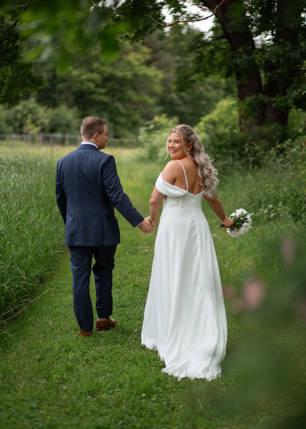 Bride and groom walk hand-in-hand through lush green garden path, bride glancing back smiling.