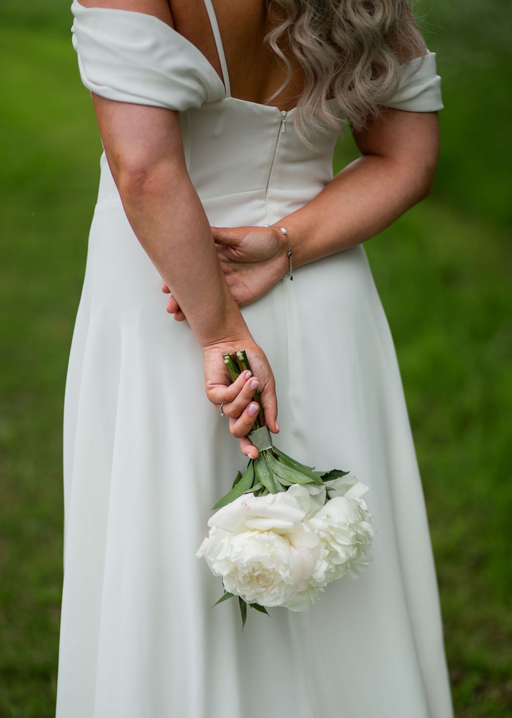 Bride in off-shoulder white gown holds white peony bouquet behind her back outdoors.