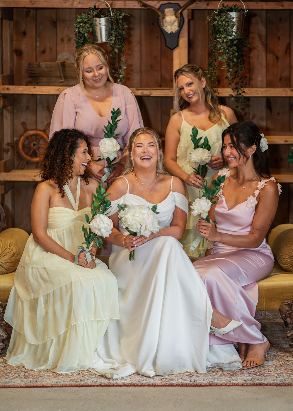 Bride in white gown laughing with four bridesmaids in pastel dresses holding white flowers in a rustic barn setting.