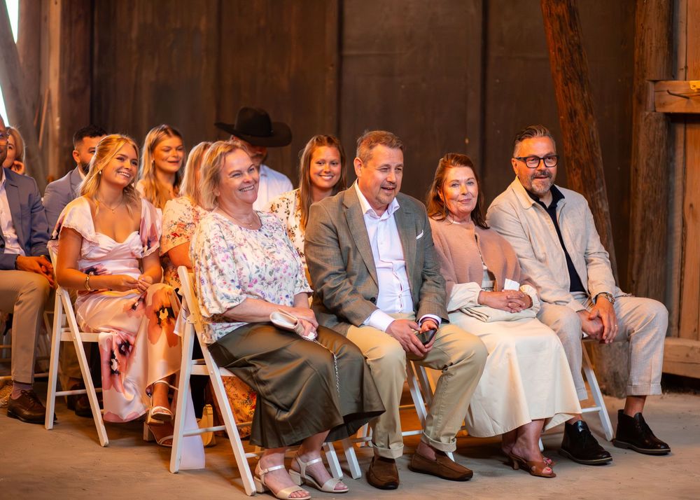 Guests smile and laugh while seated in white folding chairs during a rustic barn wedding ceremony.