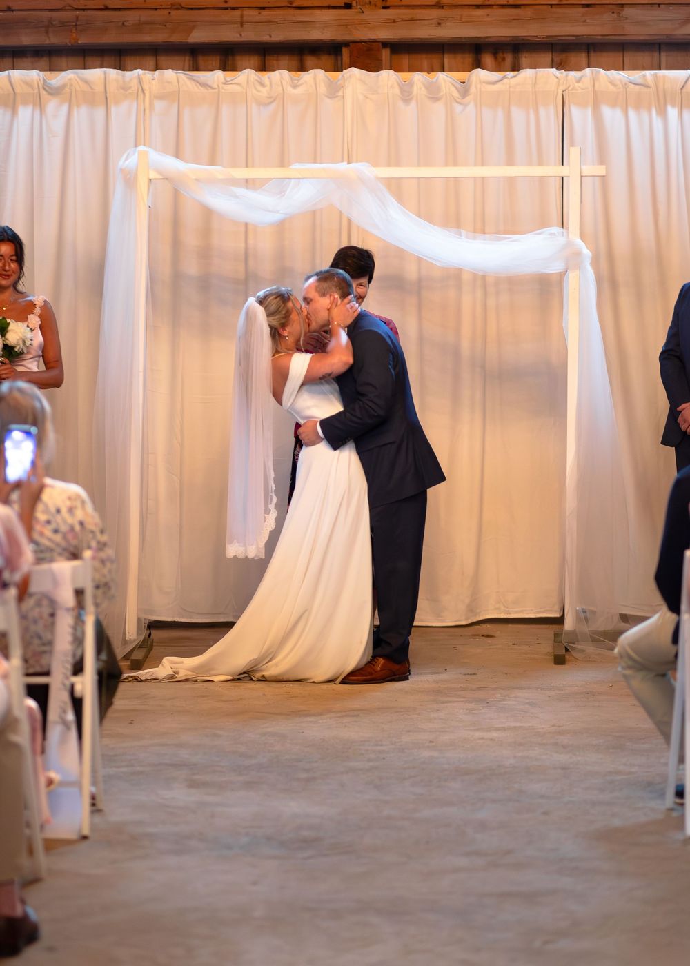 Bride and groom sharing first kiss at altar during indoor barn wedding ceremony.