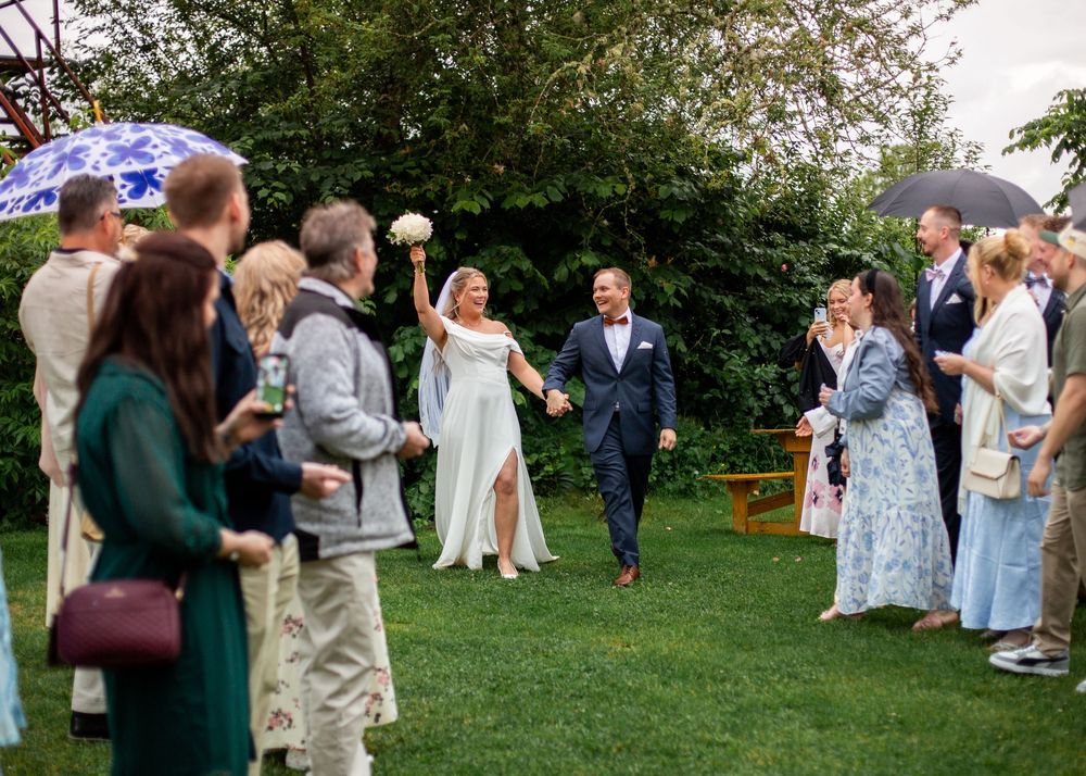 Newlyweds walk joyfully through guests outdoors as bride raises bouquet in lush garden ceremony.