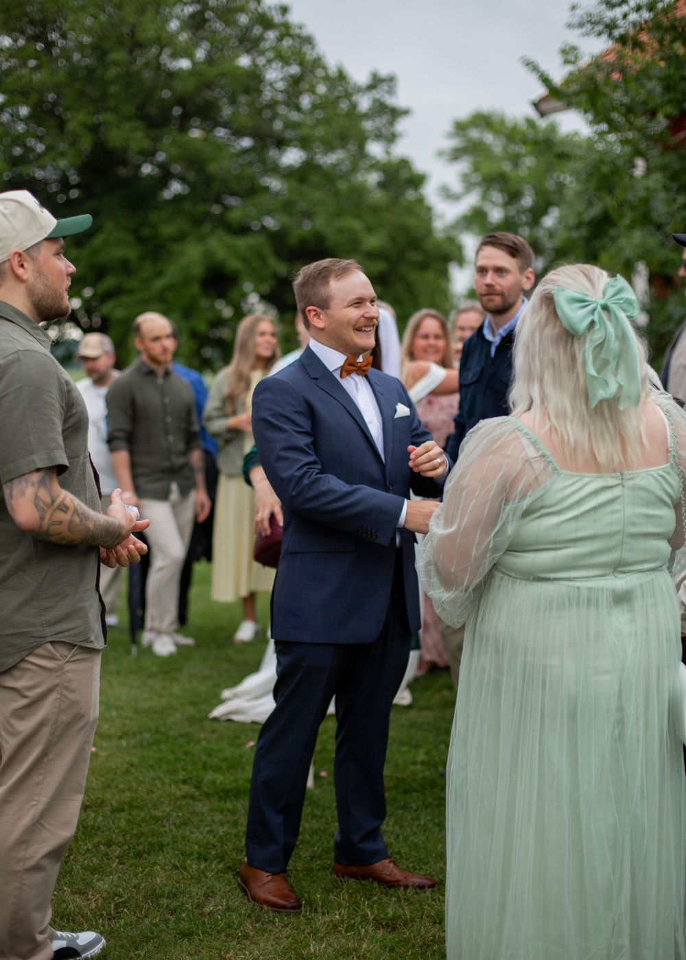A groom in a navy suit with bow tie smiles at wedding guests outdoors on a green lawn.