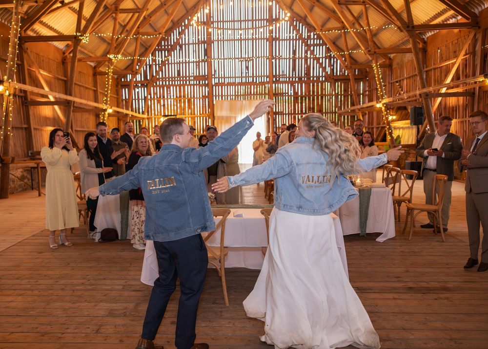 Bride and groom share first dance in matching denim jackets at a warmly lit rustic barn wedding reception.
