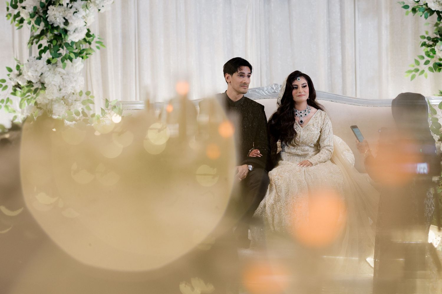 Elegant couple seated together at a wedding reception with white floral decor and warm candlelight ambiance.