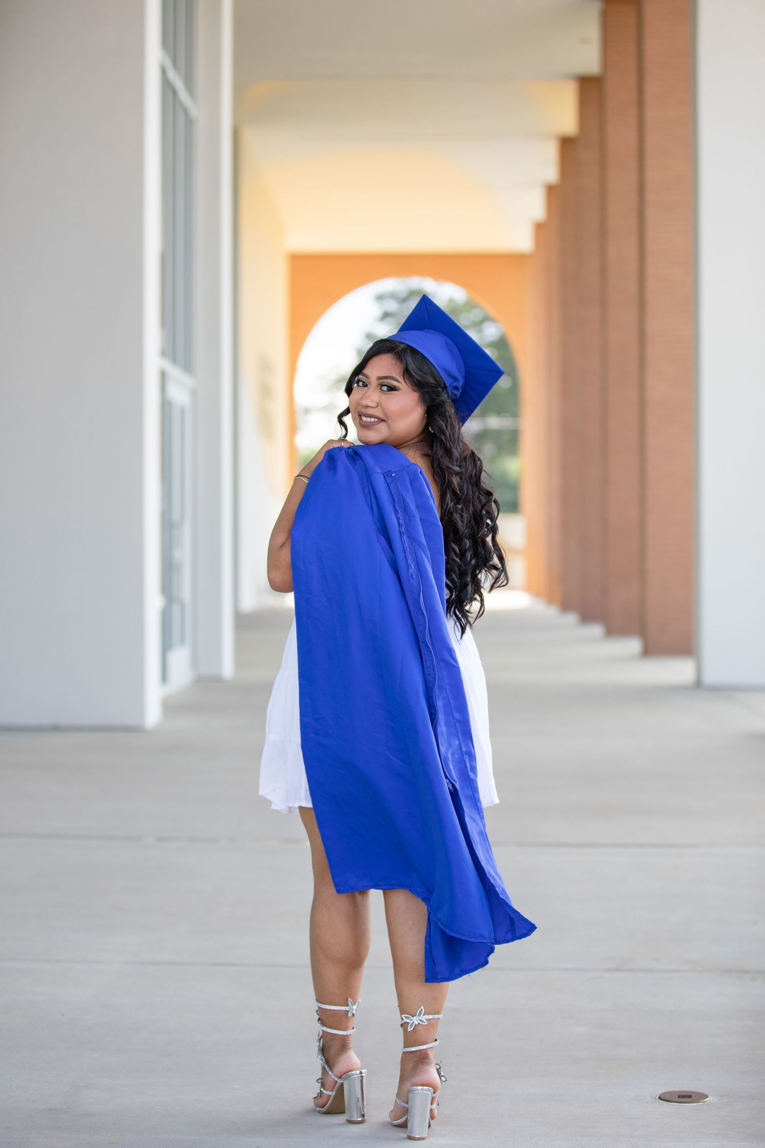 Graduate in orange cap and gown stands on wooden bridge surrounded by sunlit forest trees.