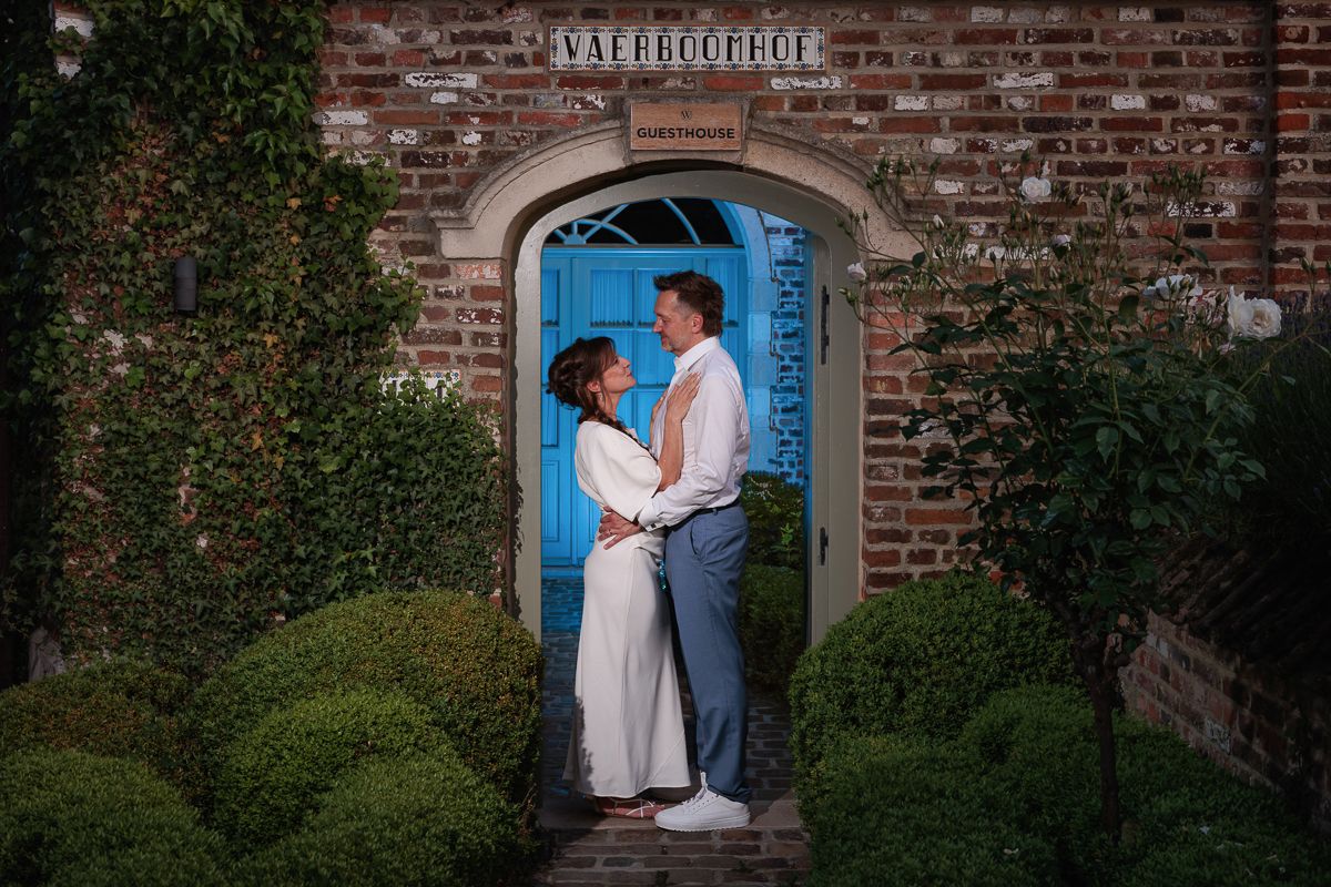 A romantic wedding couple in light gray suit and white dress share a tender moment among green foliage.