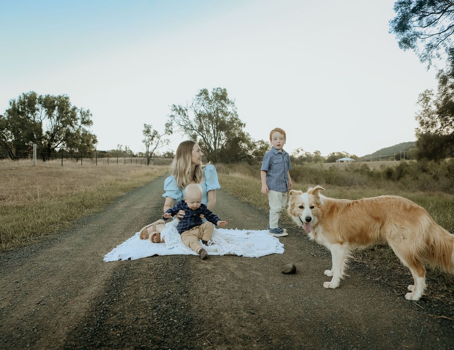 Family with two young children and a golden dog sitting on a blanket on a rural dirt road surrounded by trees.
