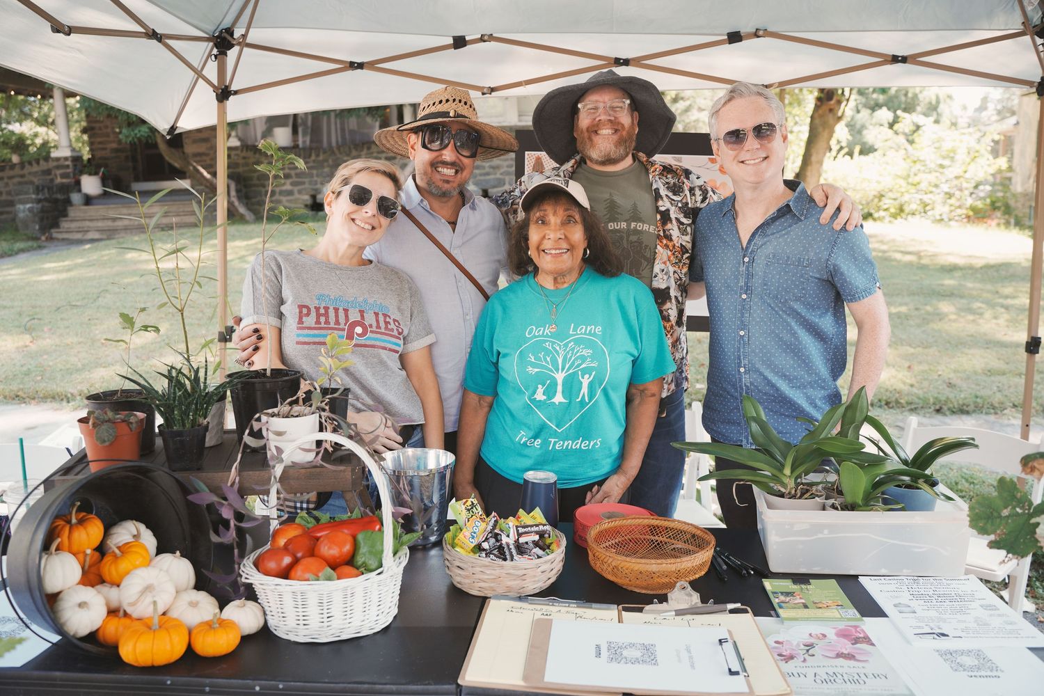 Five people smile together at an outdoor farmers market booth with fresh vegetables, plants, and baskets on display.