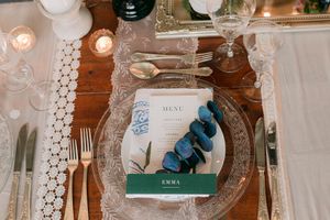 Elegant wedding table setting with gold cutlery, crystal glassware, lace runner, menu card, and blue eucalyptus sprig on a wooden table.
