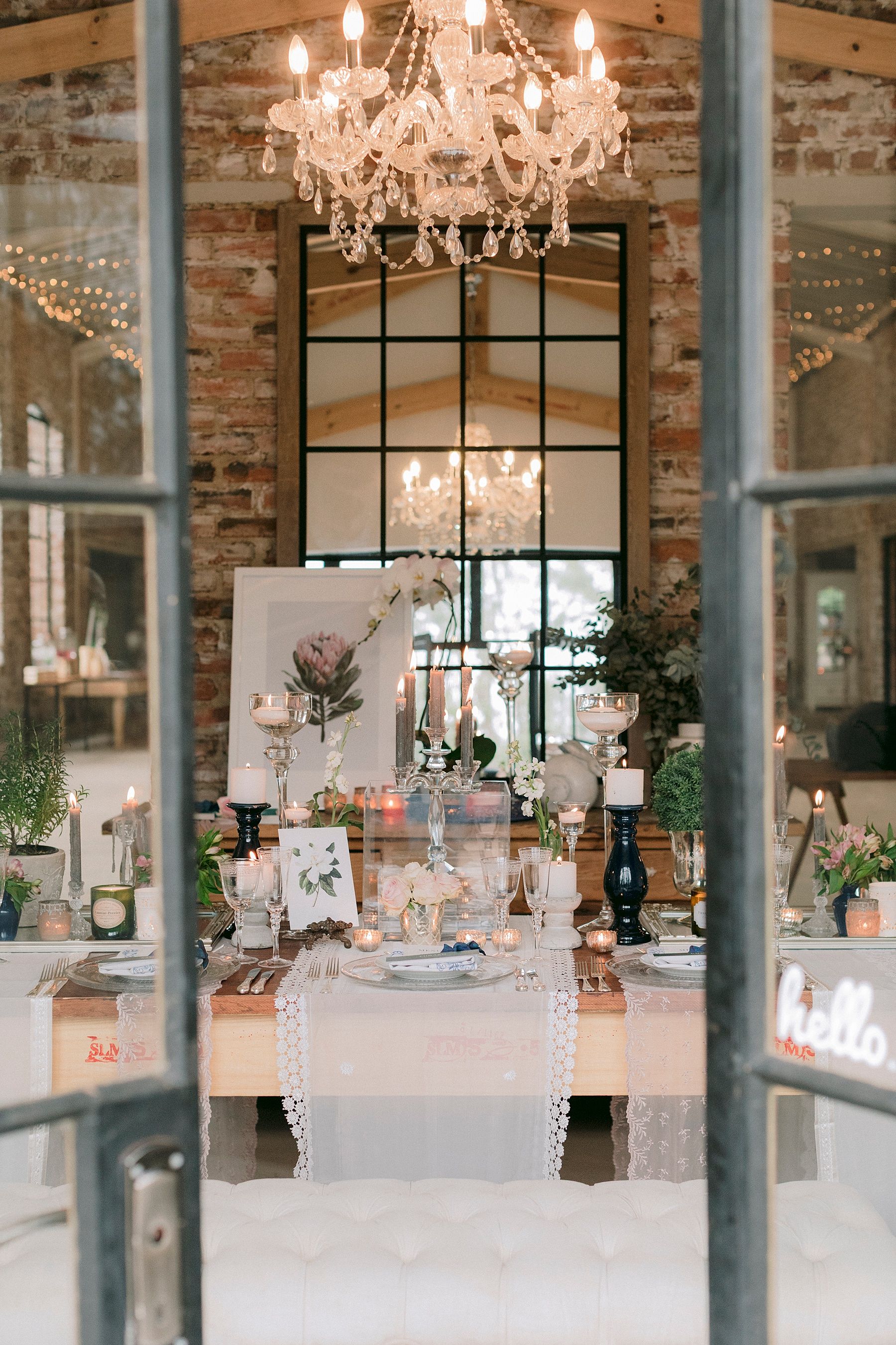 Elegant wedding reception table with chandelier, brick walls, lace runner, candles, and floral arrangements in a rustic venue.