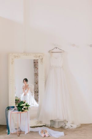 Bride in mirror reflection beside hanging white wedding gown, floral bouquet, and pink shoes in bright bridal suite.