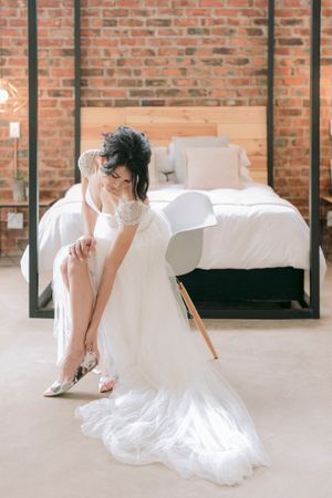 Bride in white lace gown putting on silver heels while seated on chair in rustic brick bedroom.