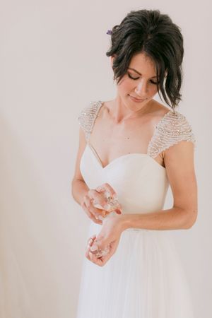 Bride in white beaded gown holding crystal jewelry, smiling softly with elegant updo hairstyle.