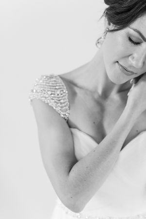 Black and white bridal portrait of a smiling bride in a beaded gown with pearl shoulder detail, eyes closed.