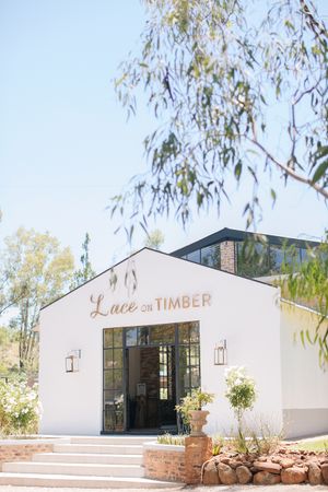 White exterior of Lace on Timber wedding venue with black-framed glass doors, brick steps, and eucalyptus trees under a clear blue sky.