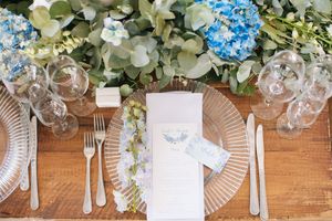 Elegant wedding table setting with blue hydrangeas, eucalyptus, glass charger plates, crystal glasses, and a menu card.