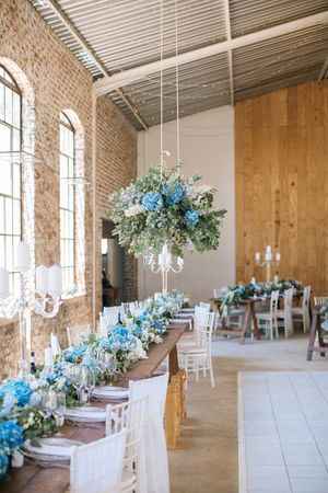 Elegant wedding reception with blue hydrangea floral arrangements on rustic wooden tables in a brick venue.