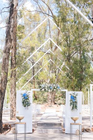 Elegant outdoor wedding ceremony setup with white geometric arch, blue floral arrangements, and tree-lined aisle.