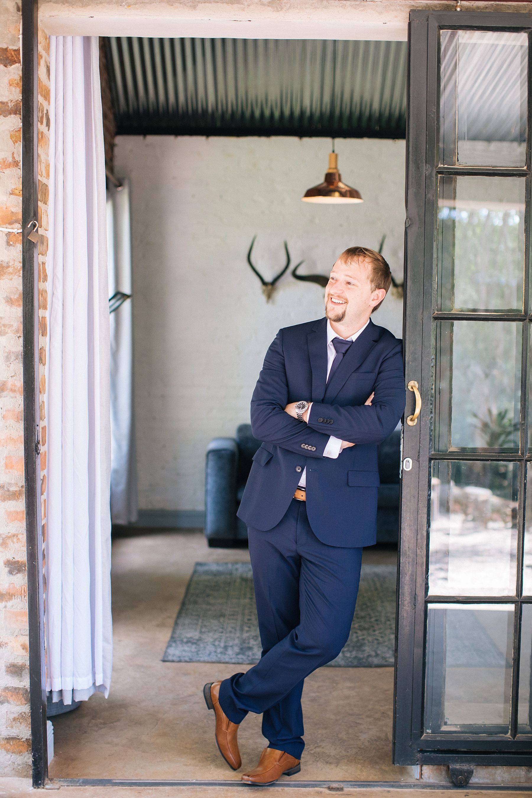 Man in navy suit leaning against open door frame with arms crossed, smiling in a rustic interior space.