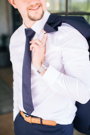 Smiling groom in white shirt, navy tie, and watch adjusting his tie before the wedding ceremony.