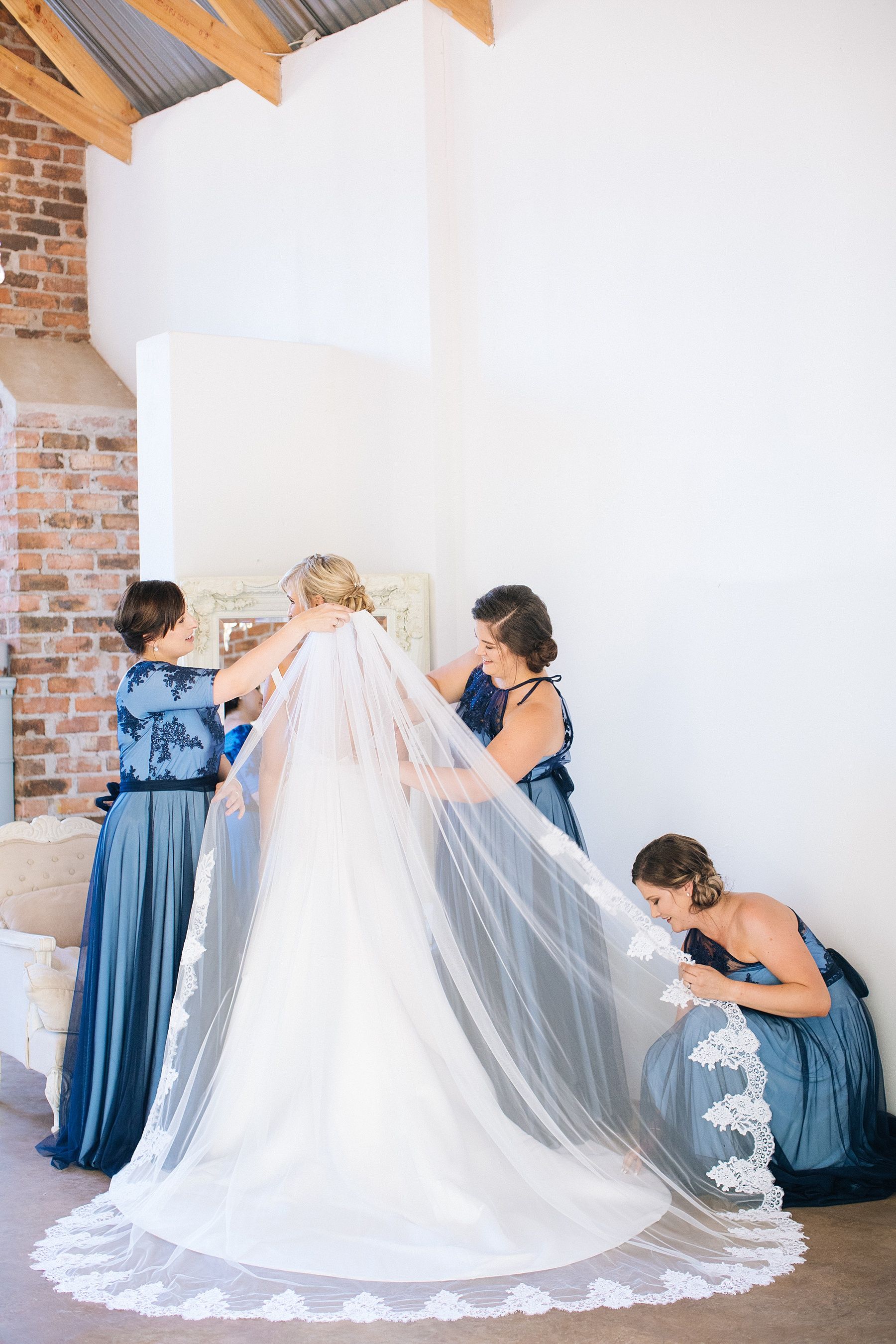 Bridesmaids in blue gowns help bride arrange her lace-trimmed cathedral veil in a bright white wedding venue.