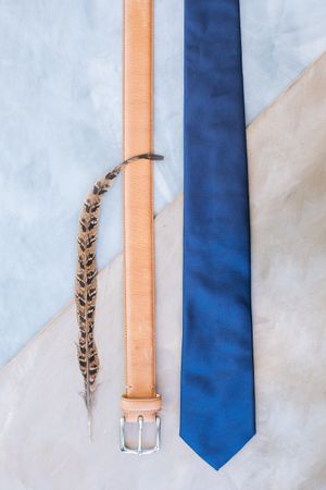 Tan leather belt, blue silk tie, and pheasant feather arranged on marble surface for a stylish men's accessory flat lay.