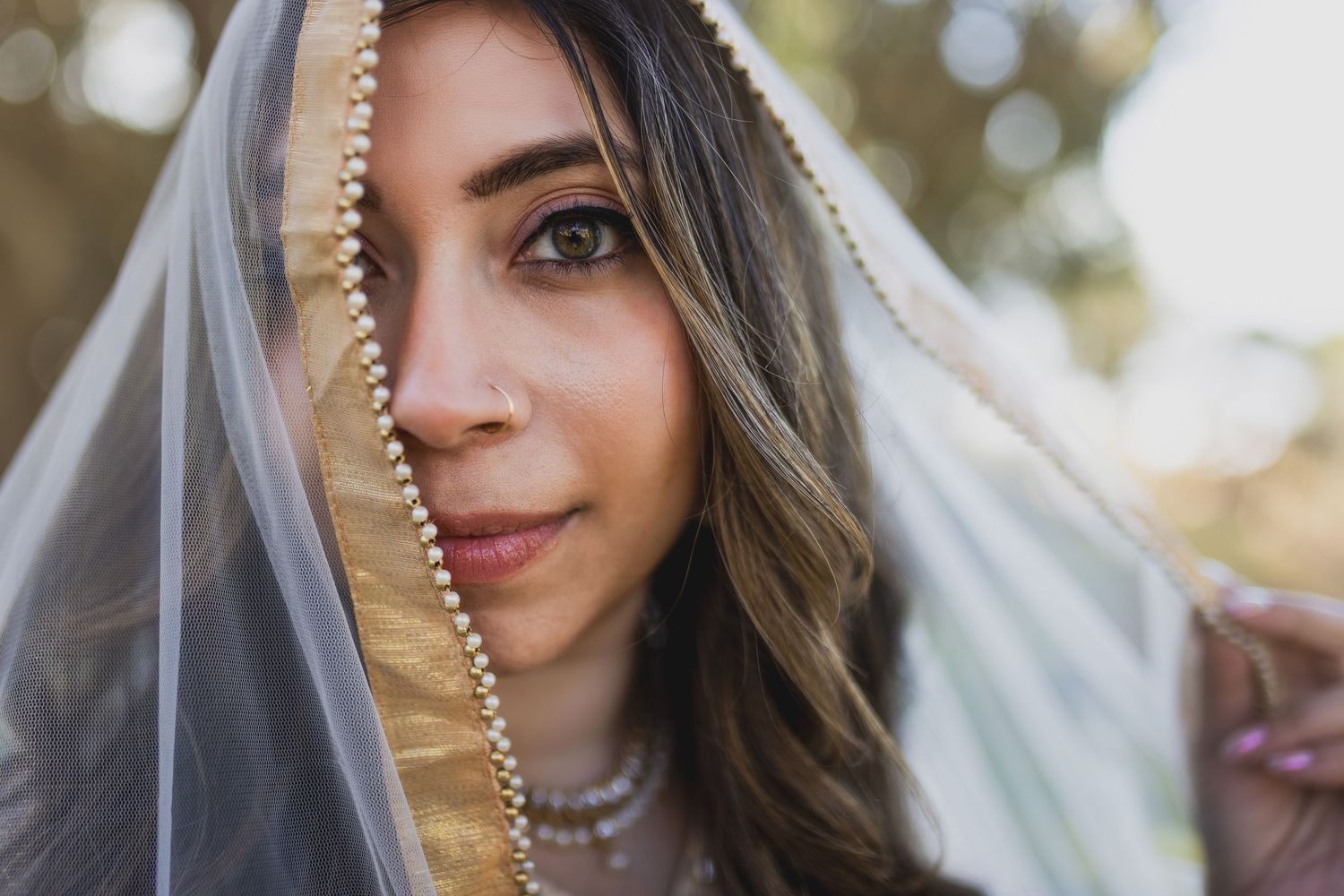 A beautiful bride with brown hair peers over a pearl-trimmed veil, her eyes gazing softly at the camera.