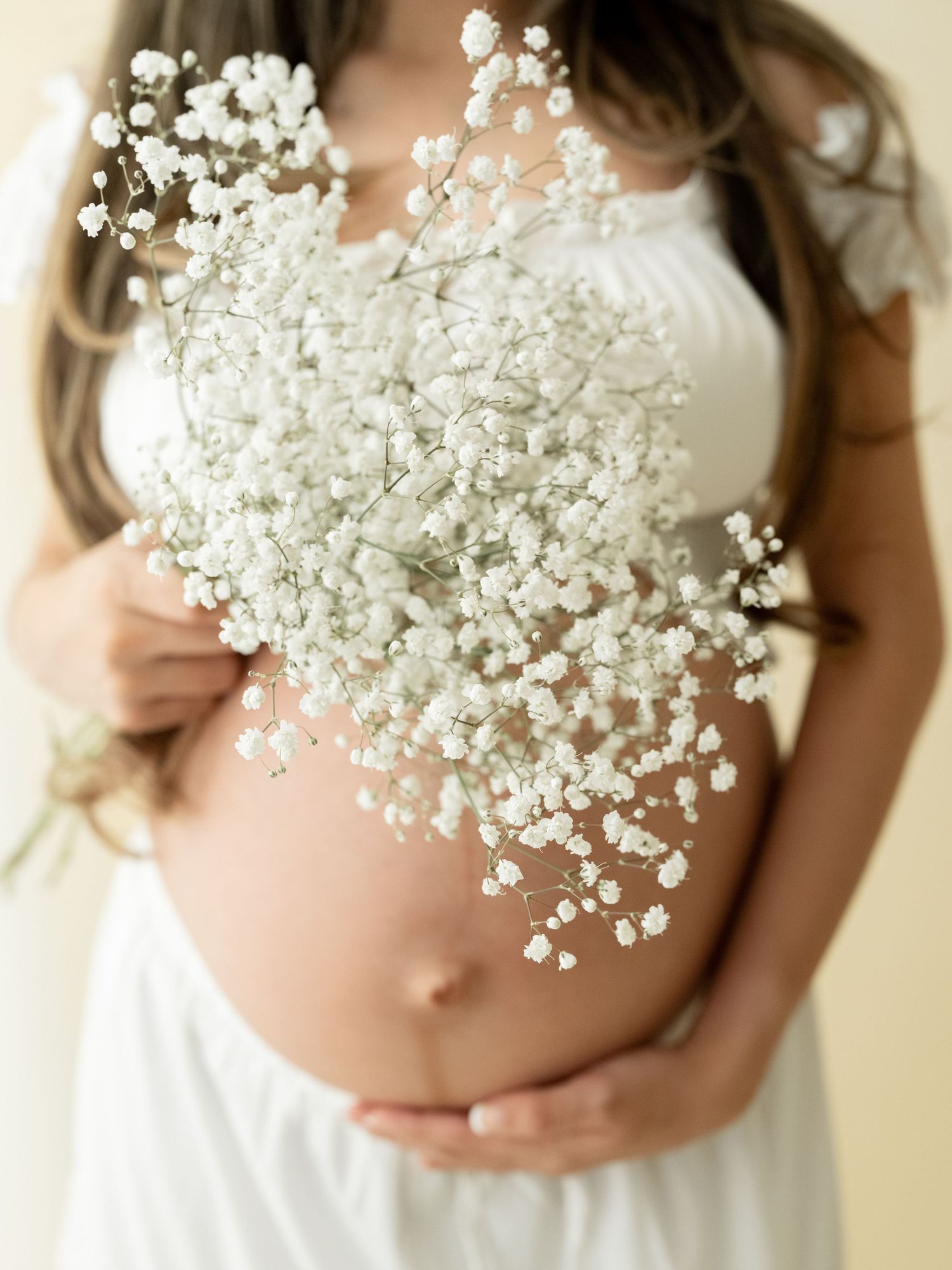 Pregnant woman in white crop top and ripped jeans cradling baby bump against white wall on wooden floor.