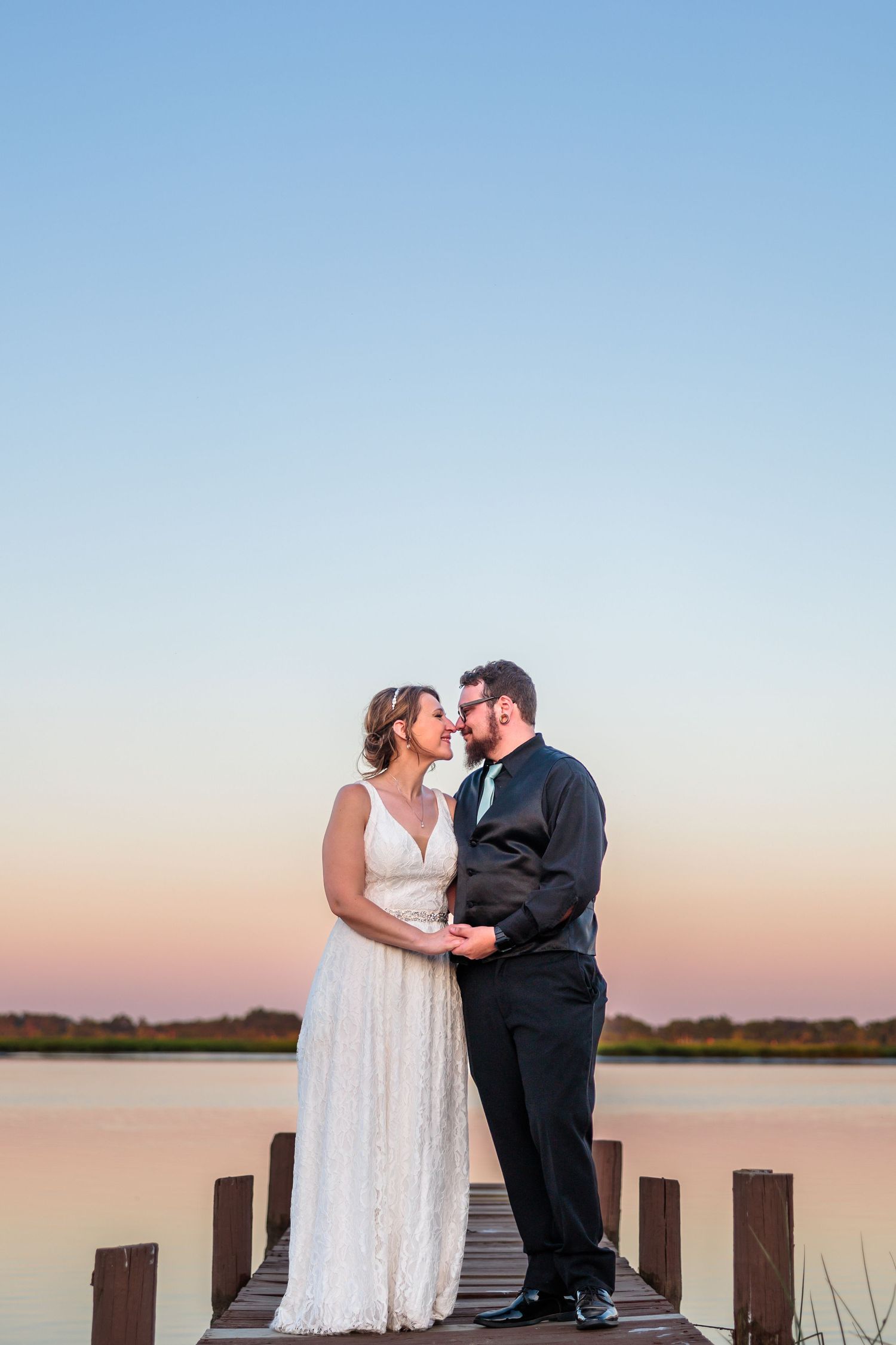 Wedding couple embracing on wooden dock during sunset over calm water.