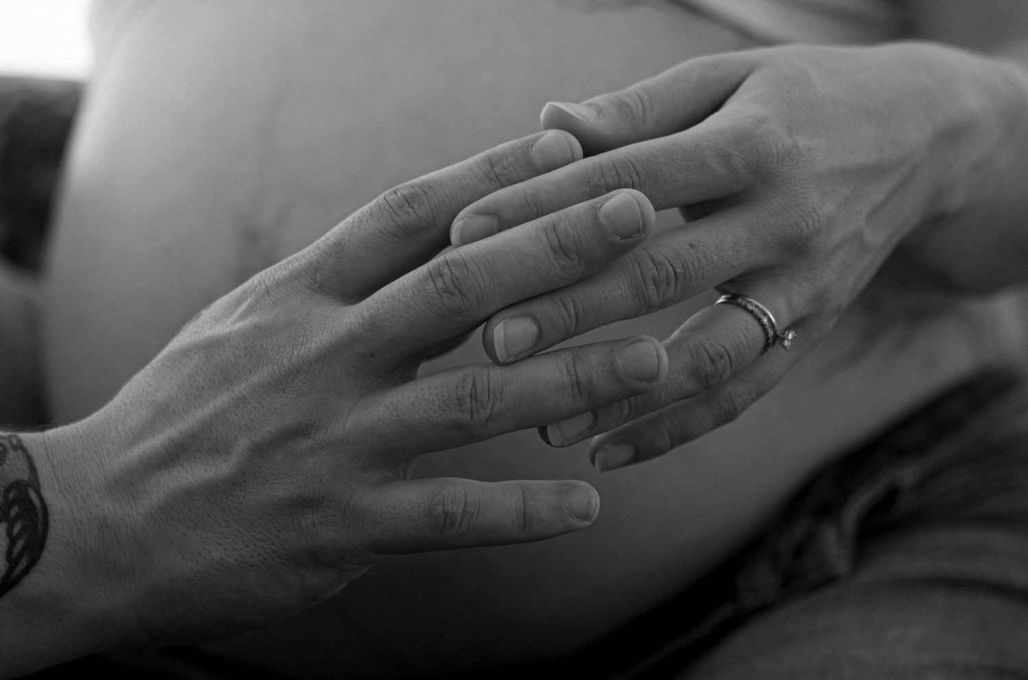 Black and white photo of two hands, one with a wedding ring, gently resting on a pregnant belly.