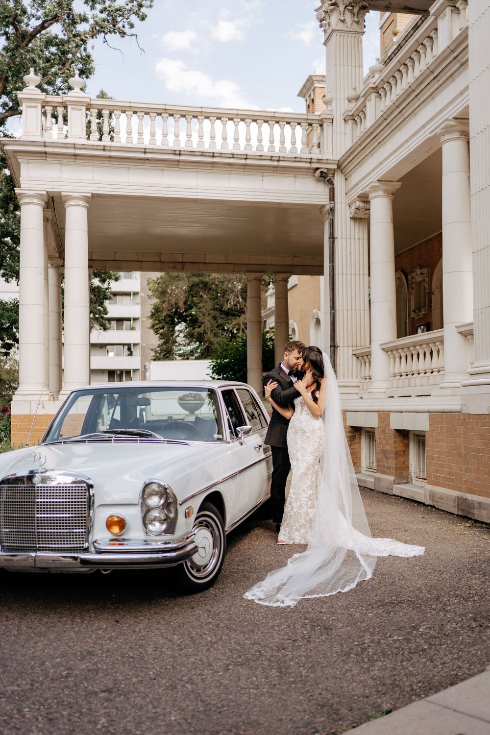Bride and groom embracing beside a vintage white Mercedes-Benz outside an elegant columned mansion.