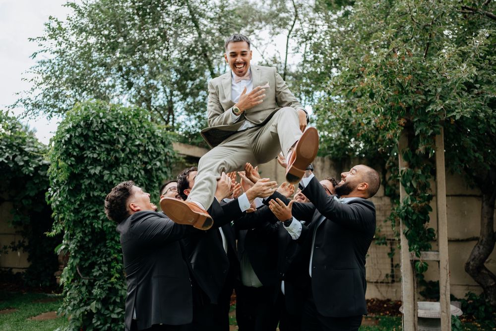 Groomsmen tossing groom in the air outdoors during a wedding celebration surrounded by lush green trees.