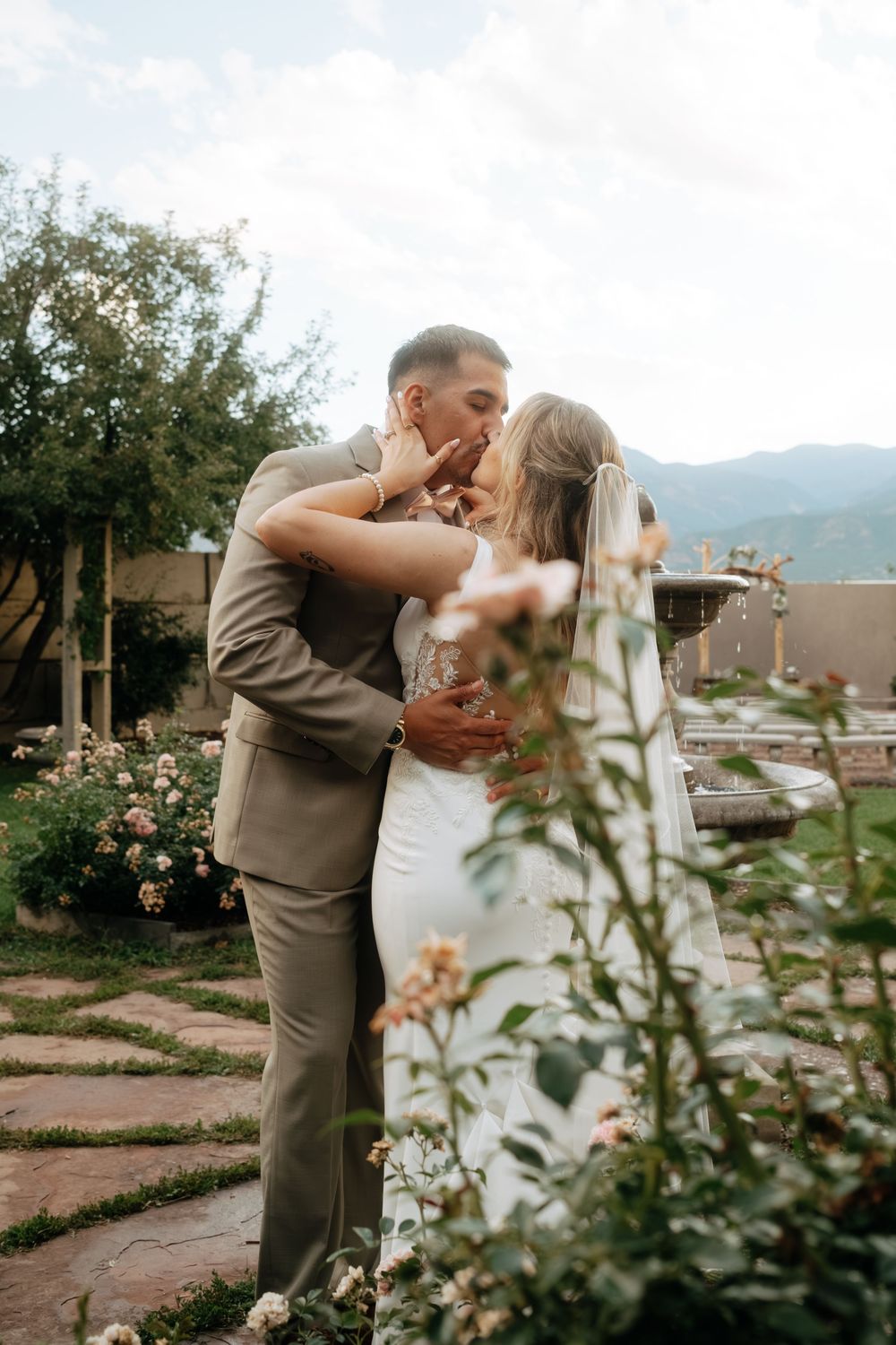 Bride and groom share a romantic kiss in a garden setting with mountain views, surrounded by blooming flowers.