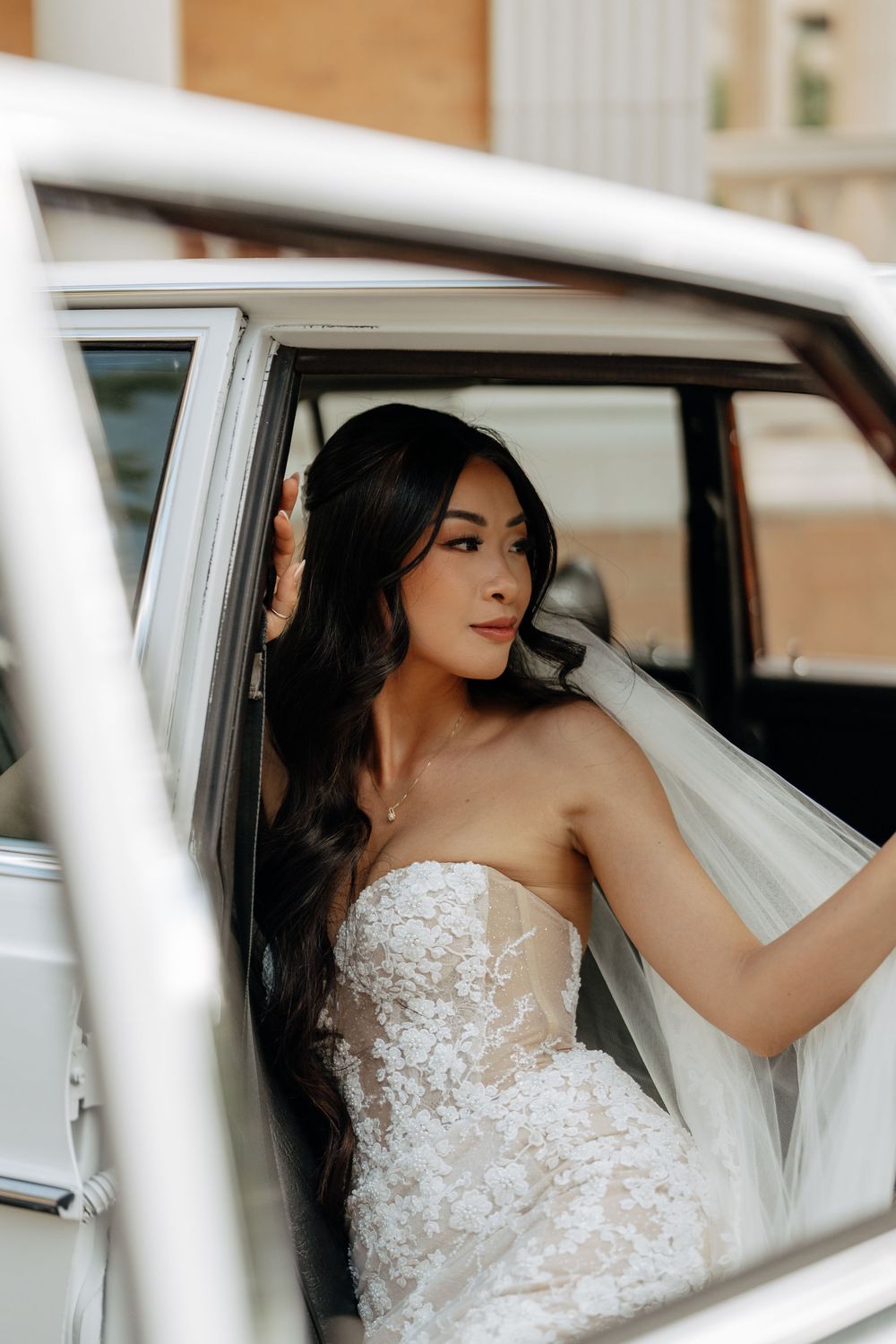 Bride in strapless lace wedding gown sitting in a vintage white car, holding her veil, with long dark hair.