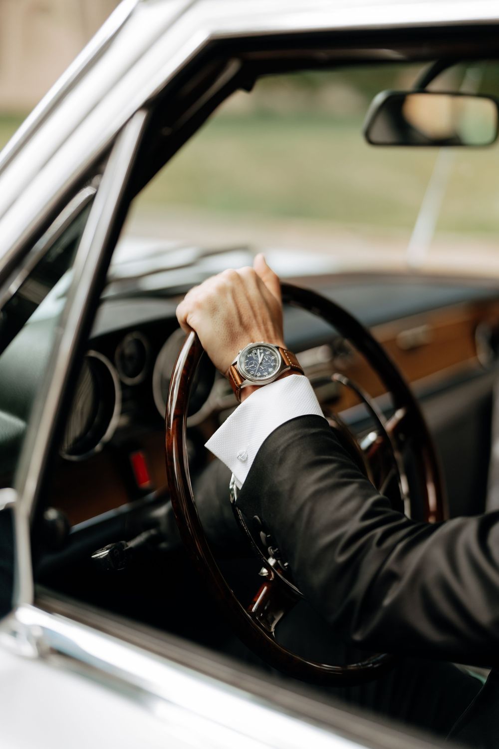 Man in suit with luxury brown watch driving a classic vintage car, close-up of hand on wooden steering wheel.