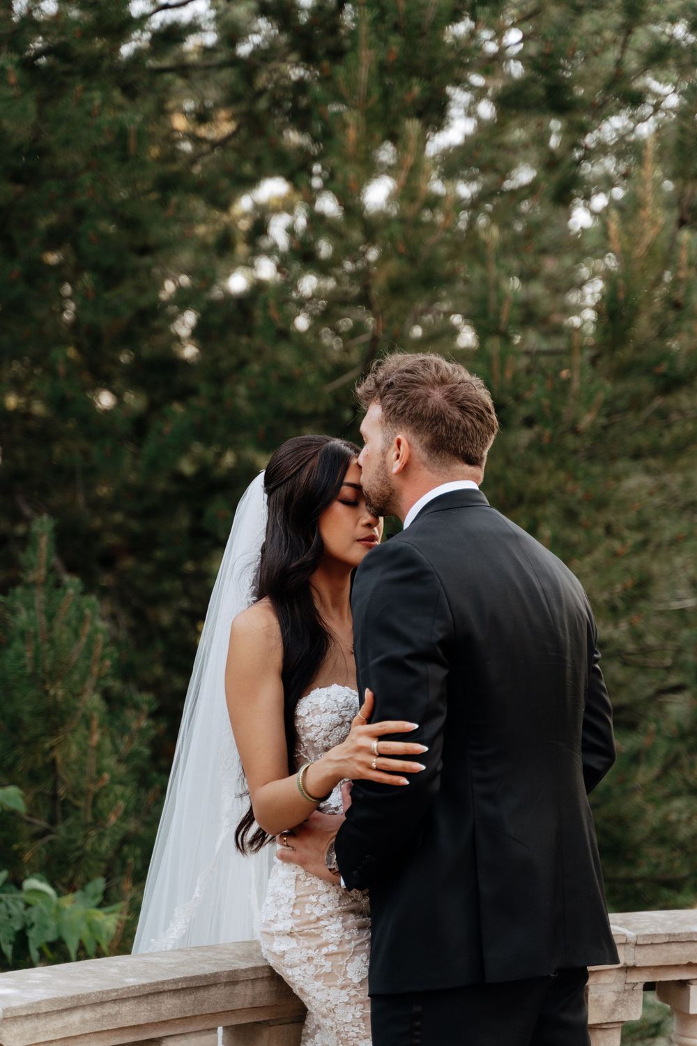 Bride and groom share a romantic kiss outdoors, surrounded by lush green trees, bride wearing lace gown and veil.