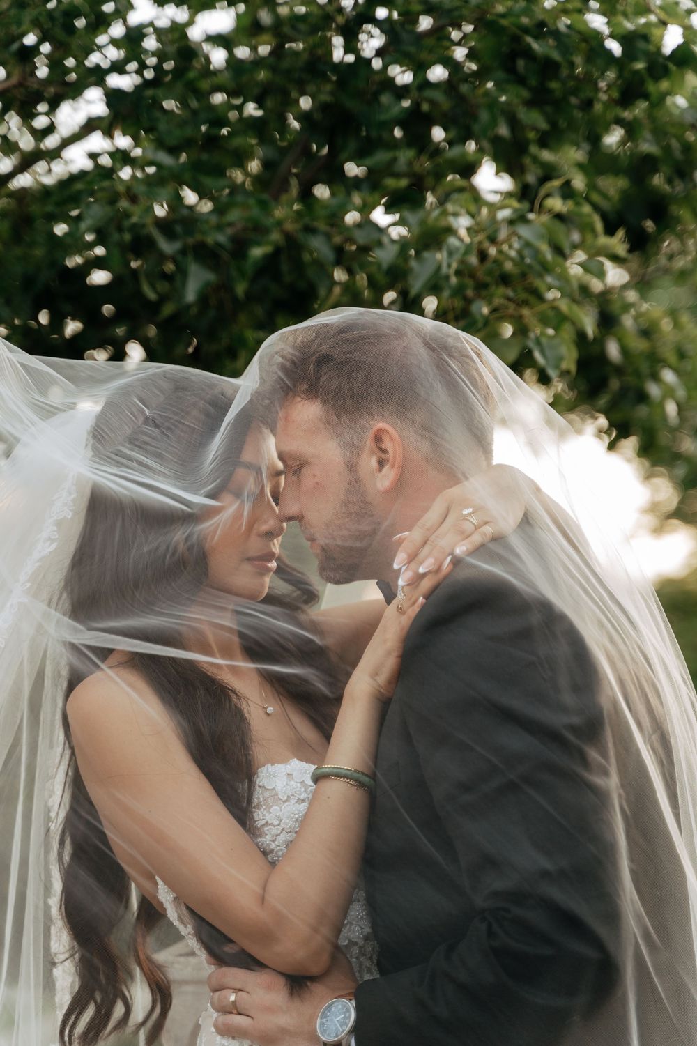 Bride and groom share a romantic moment under a flowing white veil outdoors, surrounded by lush green trees.