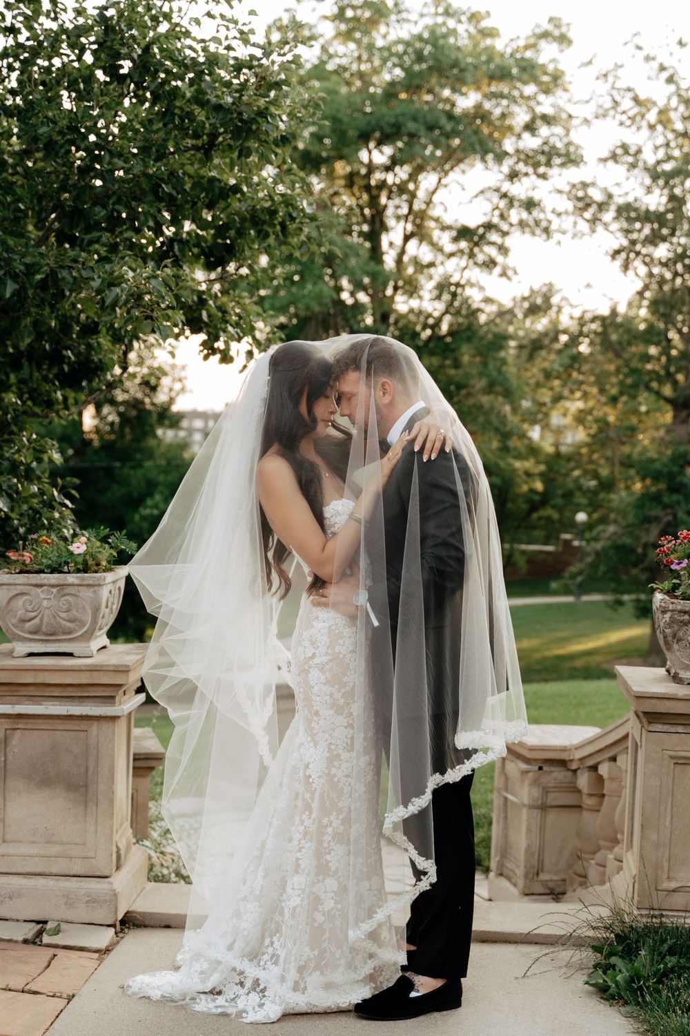 Bride and groom share a romantic moment under a flowing lace wedding veil in a lush garden setting.