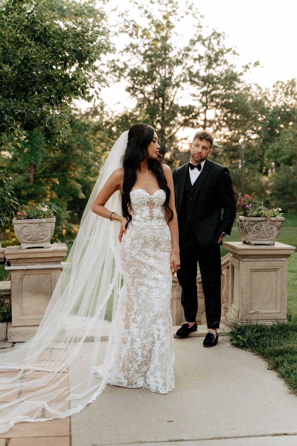 Bride in lace gown with veil and groom in black tuxedo standing together on stone terrace surrounded by lush greenery.
