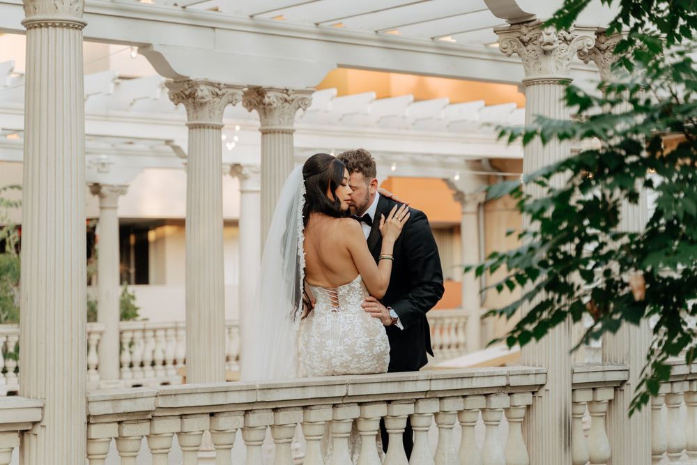 Bride and groom share romantic kiss on elegant white balcony with columns on wedding day.
