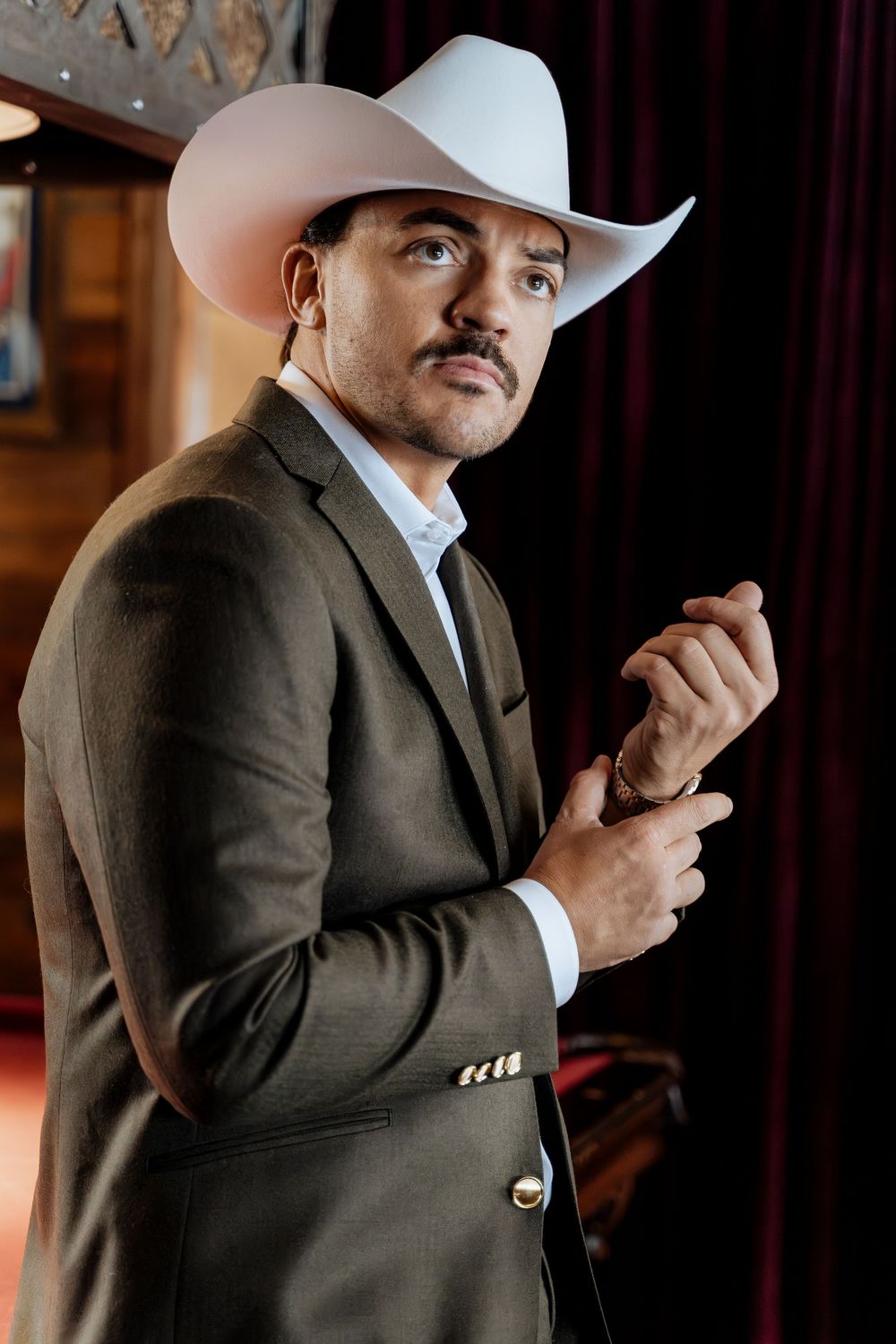 Man in white cowboy hat and dark suit adjusting cufflinks, standing in dimly lit venue.