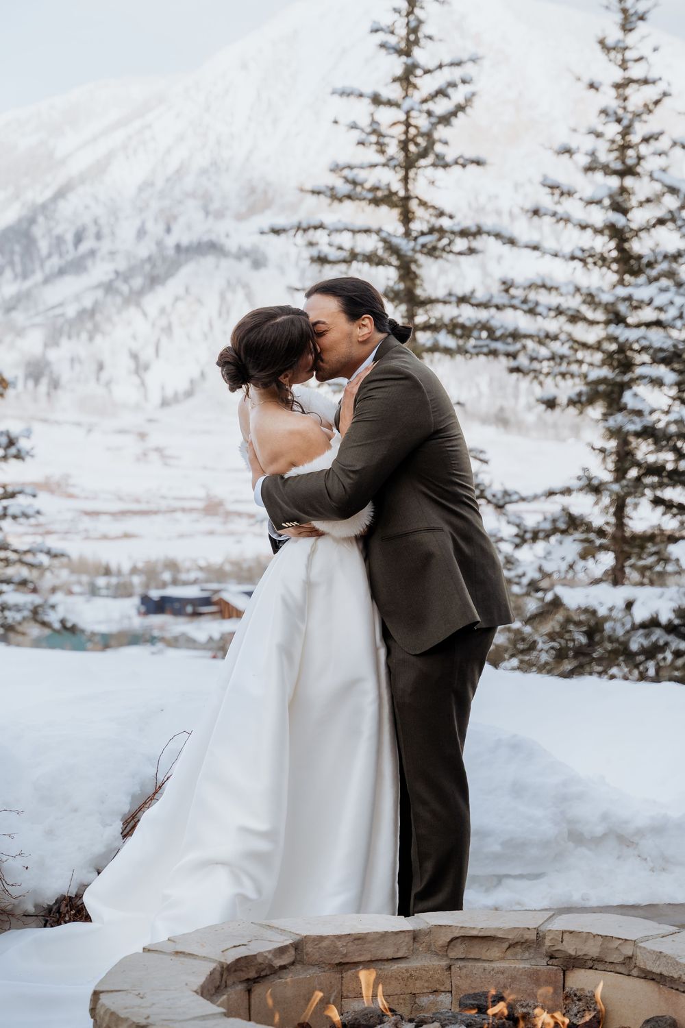 Couple sharing a romantic kiss in wedding attire beside a stone fire pit with snowy mountains and pine trees behind them.