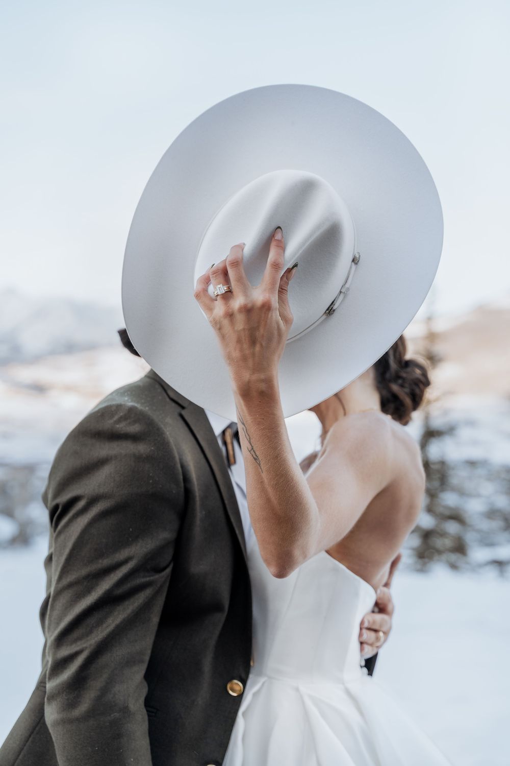 Bride and groom kissing behind a wide-brim white hat in a snowy mountain wedding setting.