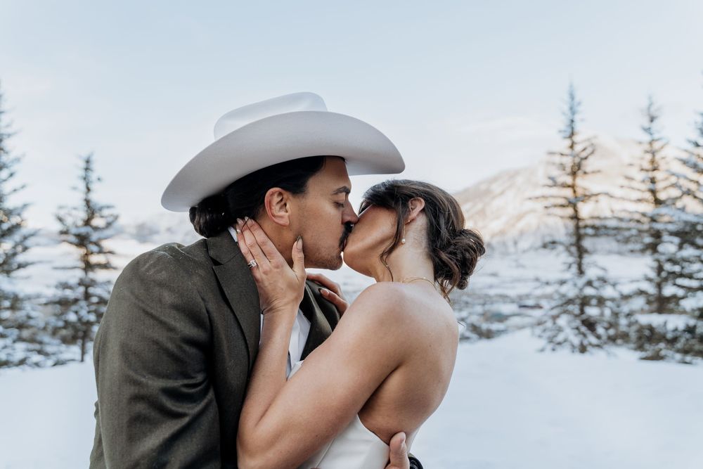 Bride and groom sharing a romantic kiss in a snowy mountain setting, groom wearing a white cowboy hat and grey suit.