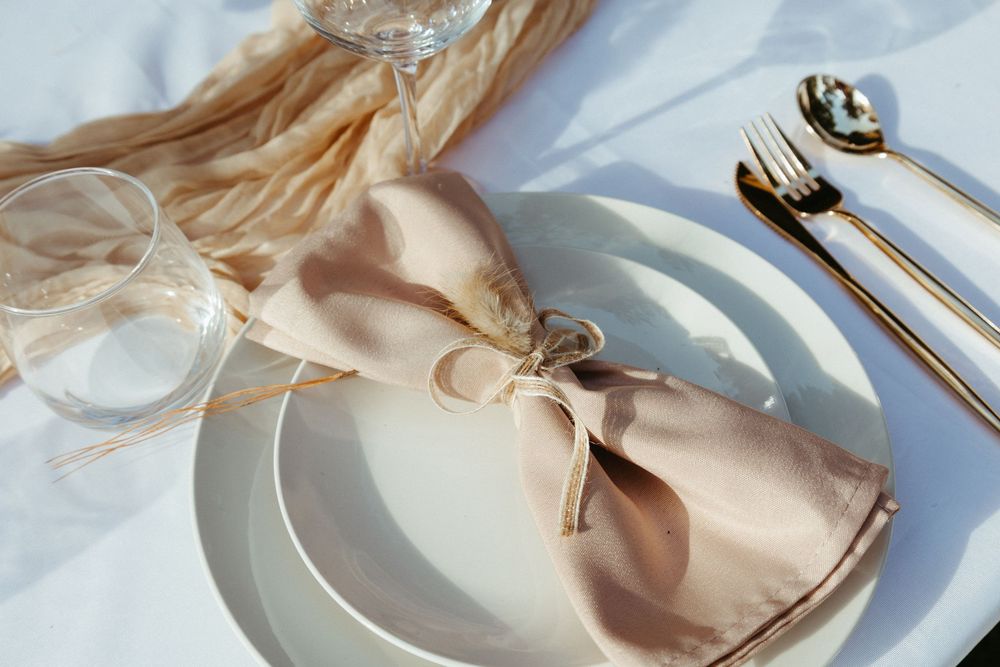 Elegant wedding place setting with blush napkin bow tie, white plates, gold cutlery, and crystal glassware.