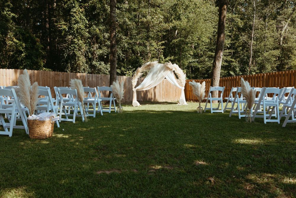 Outdoor wedding ceremony setup with white chairs, pampas grass arch, and lush green lawn surrounded by trees.
