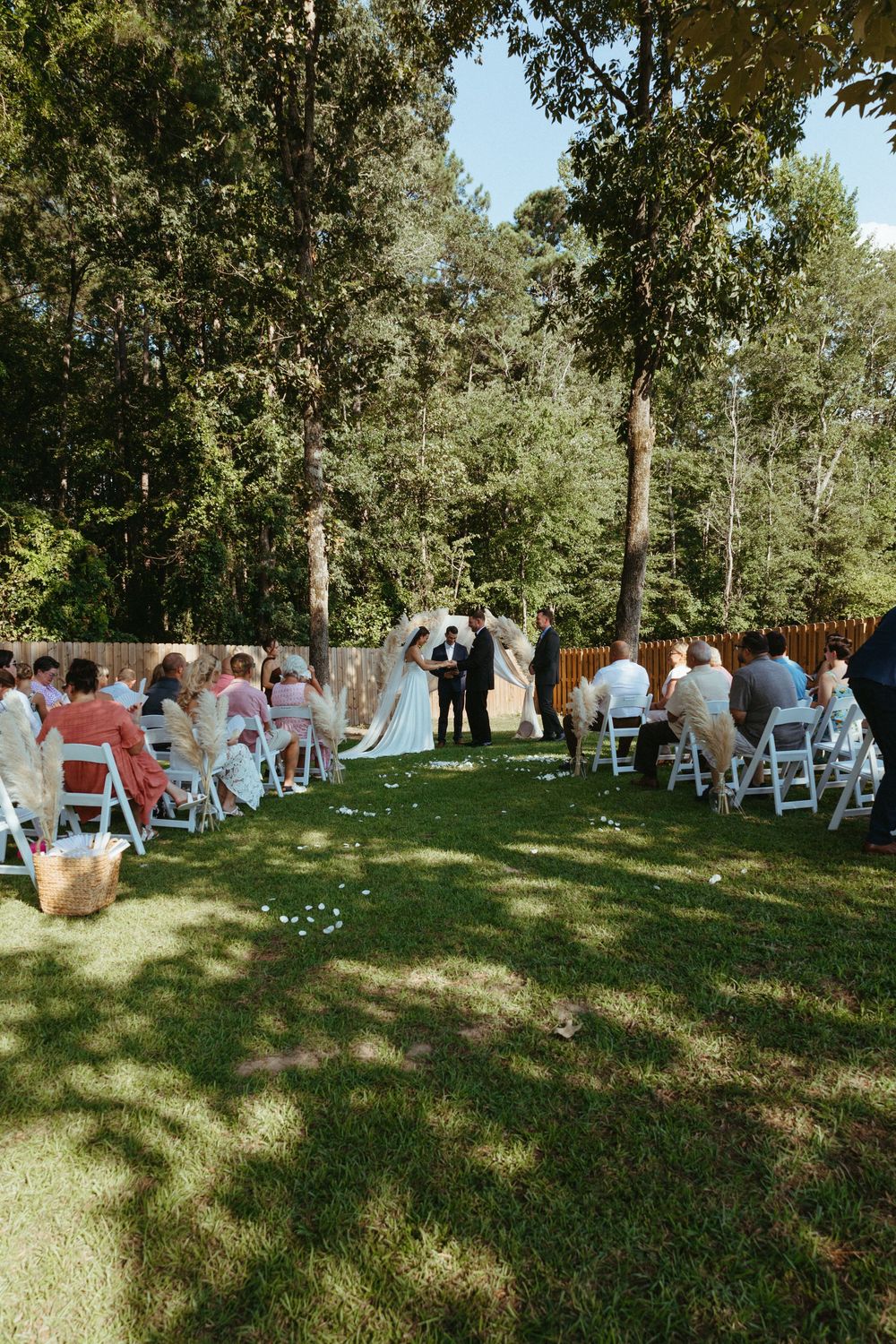 Outdoor wedding ceremony on green lawn with white chairs, couple at altar, surrounded by tall trees and wooden fence.